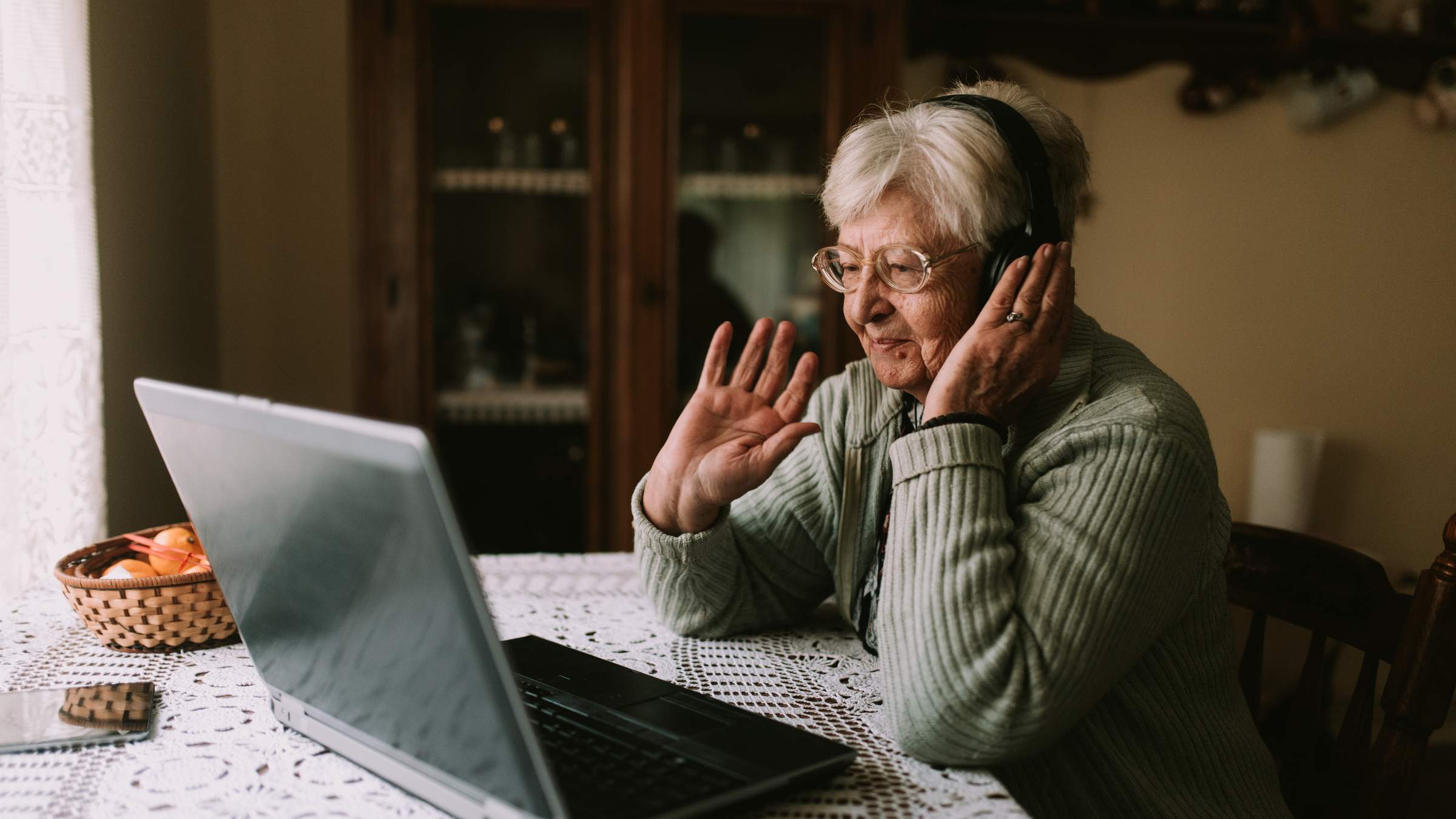 Elderly woman with headphones smiling and waving at her laptop screen during a video call at her kitchen table